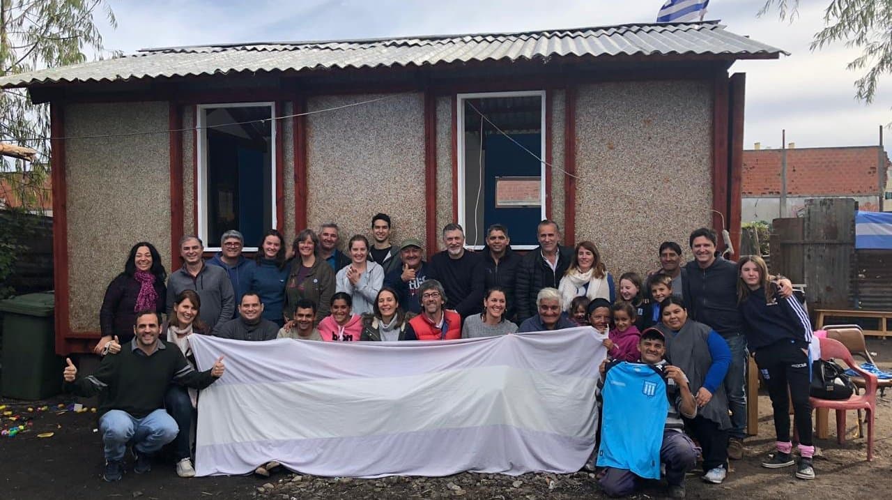 Gente reunida con una gran bandera frente a una pequeña vivienda.
